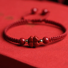 Red braided cord bracelet with cinnabar four-leaf clover charm, gold accent beads and round red stone beads on matching red fabric background