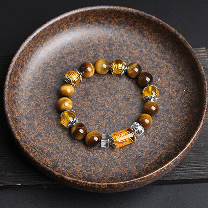 Overhead view of tiger eye gemstone bracelet with carved amber cylindrical focal bead, ornate silver-toned caps and faceted crystal spacers in round ceramic dish on dark wood
