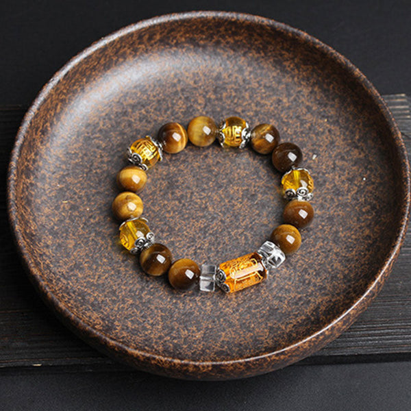 Overhead view of tiger eye gemstone bracelet with carved amber cylindrical focal bead, ornate silver-toned caps and faceted crystal spacers in round ceramic dish on dark wood