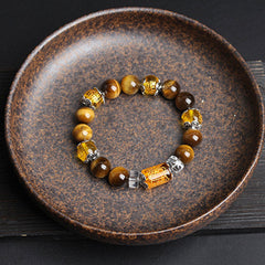Overhead view of tiger eye gemstone bracelet with carved amber cylindrical focal bead, ornate silver-toned caps and faceted crystal spacers in round ceramic dish on dark wood