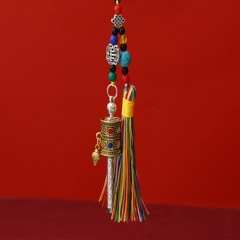 Tibetan Prayer Wheel Keychain – two-tone silver and gold prayer wheel pendant with colorful silk tassel, turquoise beads and auspicious knot charm, red background, front vertical view