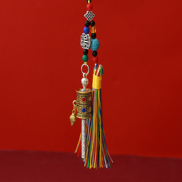 Tibetan Prayer Wheel Keychain – two-tone silver and gold prayer wheel pendant with colorful silk tassel, turquoise beads and auspicious knot charm, red background, front vertical view