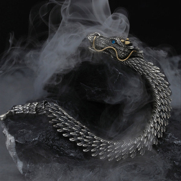 Dragon cuff bracelet with articulated scale segments blue gemstone eye gold whiskers and antiqued silver finish displayed in dynamic S-curve on dark stone with mist effects