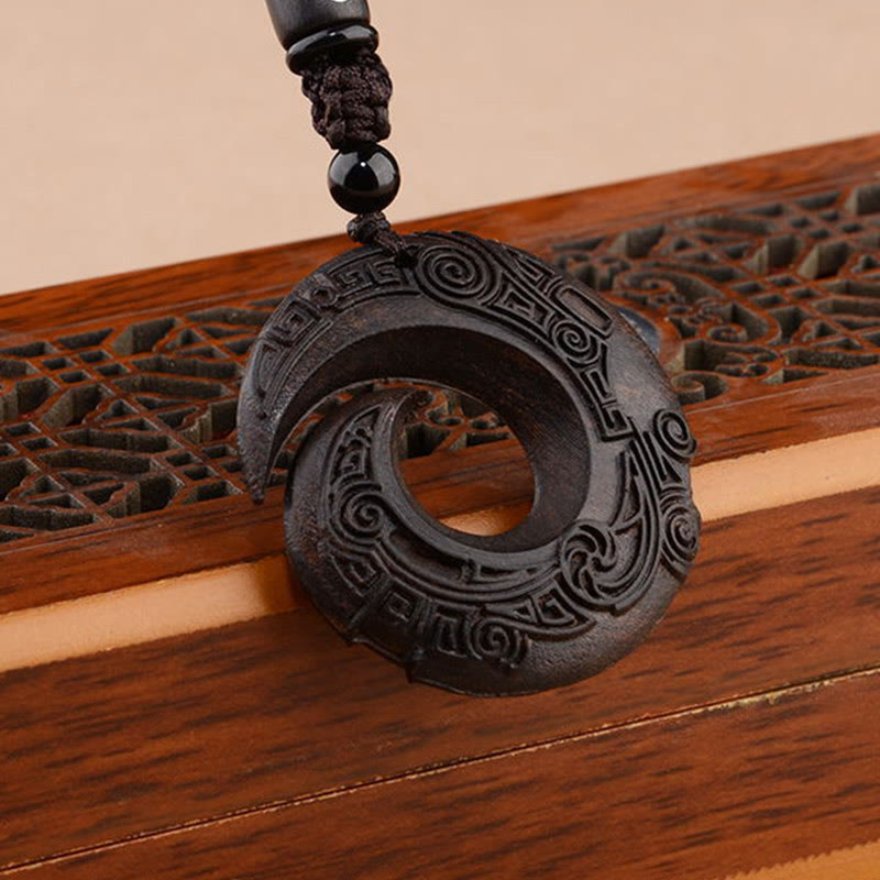 Dark sandalwood peace buckle pendant displayed on carved wooden lattice showing interlocking double-ring eternity spiral design with intricate relief carving and black braided cord accent