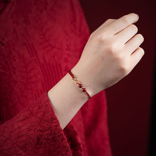 Cinnabar Red String 14K Gold Infinity Bracelet worn on wrist, burgundy silk cord with gold infinity knot connector, two deep red cinnabar beads, traditional Chinese brocade clothing, lifestyle model shot