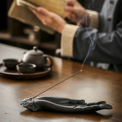 Buddha Hand Incense Burner – matte black ceramic hand holding lit incense stick, smoke rising over blurred tea set, mindful
