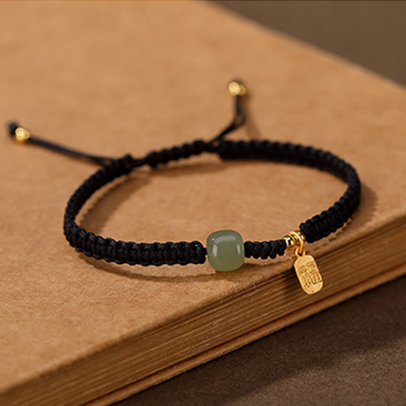 Black braided cord bracelet with Hetian jade barrel-shaped green stone bead gold fu blessing charm and gold-tipped cord ends on vintage books display with neutral gray backdrop