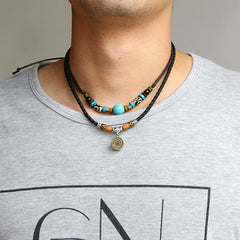 Turquoise Dzi Bead Protection Necklace – worn by male model on gray t-shirt showing double-strand design, turquoise focal bead on upper strand and antique brass coin pendant on lower strand, black braided leather cord with adjustable knot closure