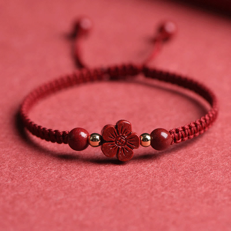 Adjustable red braided cord bracelet with carved cinnabar five-petal flower charm, gold-tone spacer beads and round red beads on textured red surface