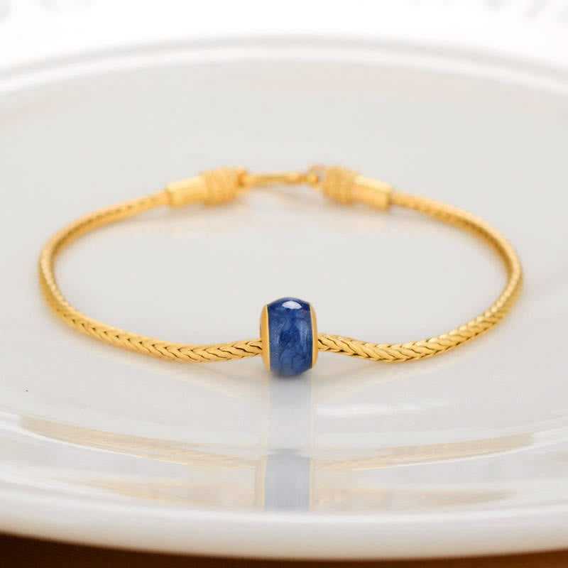 18K Gold Om Mani Padme Hum Koi Fish Bracelet – gold braided chain with blue lapis lazuli barrel bead, white ceramic dish, front-facing