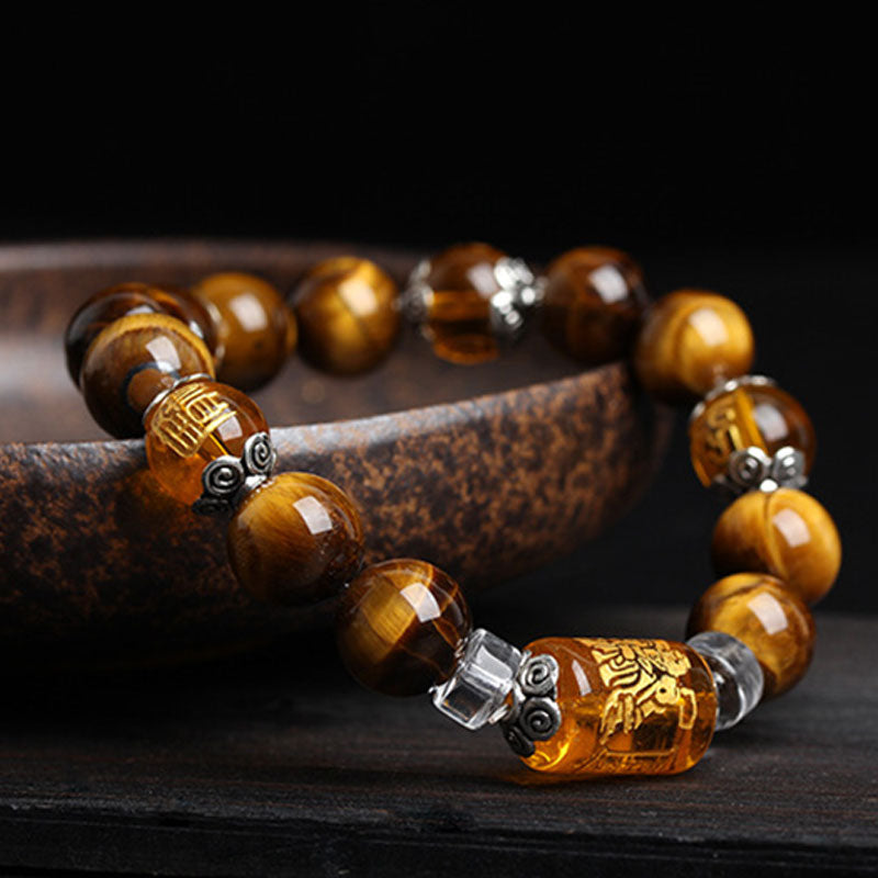 Tiger eye bead bracelet with amber barrel bead featuring deity carving, silver scroll-pattern spacers and clear crystal accents on dark wood surface with mottled ceramic dish