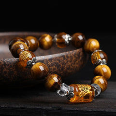 Tiger eye bead bracelet with amber barrel bead featuring deity carving, silver scroll-pattern spacers and clear crystal accents on dark wood surface with mottled ceramic dish
