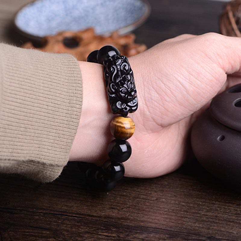 Obsidian PiXiu carved pendant close-up on dark wood, amber tiger eye bead, mythical beast detail, healing