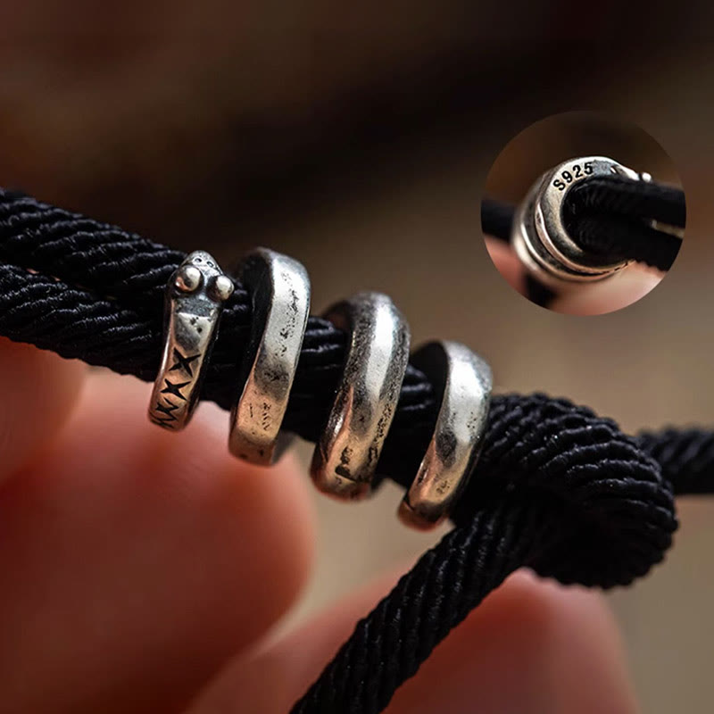 Tibetan Om Mani Padme Hum Wrap Bracelet – Black braided cord with silver engraved bead and colorful threads, coiled on beige fabric overhead view