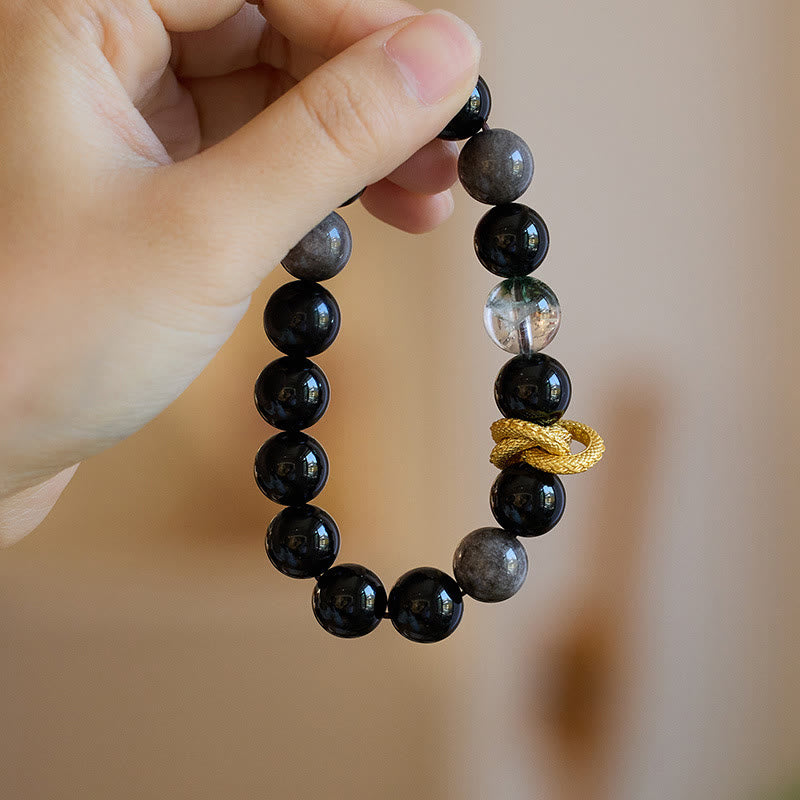 Silver Sheen Obsidian Dragon Bracelet – Hand-held view with warm bokeh background, black beads and dragon, statement
