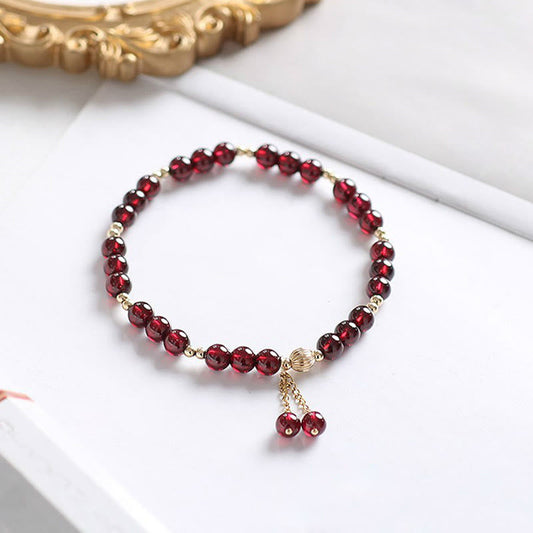 Garnet bracelet with textured gold focal bead and dual red bead dangle tassel, adjustable chain detail on white marble surface with gold baroque frame accent