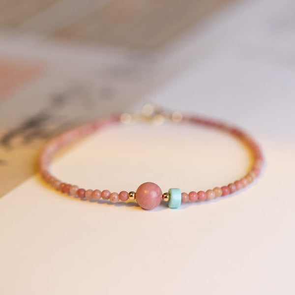Rhodonite Loving Energy Bracelet – Pink-to-coral stone beads with gold clasp hardware and turquoise accent on white surface with calligraphy corner detail