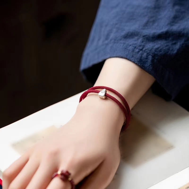 925 Silver Fu Character Braided Bracelet – Red cord bracelet with silver fortune charm on textured red circular mat with gold border, dark maroon background
