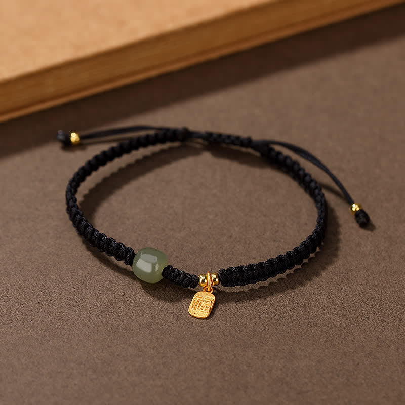 Black cord bracelet worn on model wrist showing Hetian jade green bead gold fu blessing charm pendant against chocolate brown background with cream knit sweater styling