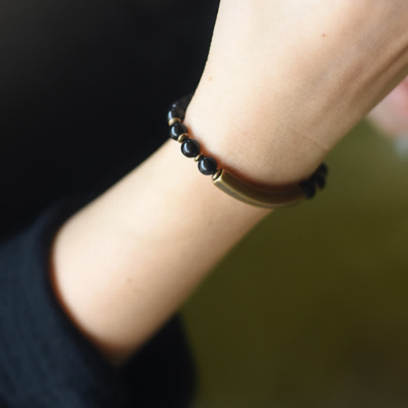 Black onyx bracelet worn on wrist with dark clothing background, polished black gemstone beads with antique bronze focal bar and metal accents lifestyle shot