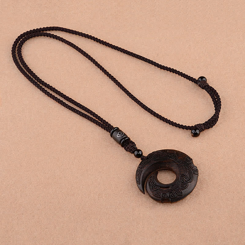 Ebony wood peace buckle pendant necklace showing circular bi-disc with spiral rotational cut and carved ancient Chinese patterns on dark wood with adjustable braided cord and cloud motif bead