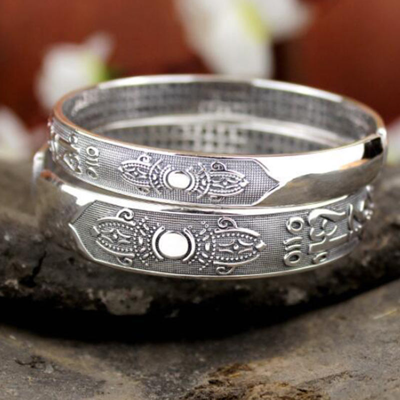 Tibetan silver prayer bangle with lotus floral medallion panels, Tibetan script, polished circular center elements, and stippled texture on dark stone with flowers.