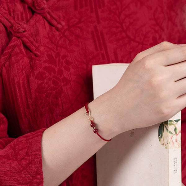 Cinnabar Red String 14K Gold Infinity Bracelet worn on wrist with Chinese brocade, braided red cord with gold connector between two cinnabar beads, sliding knot closure, traditional cultural styling with botanical book prop