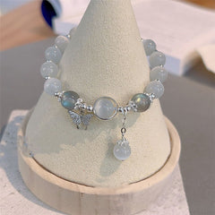 Cat's Eye Moonstone Butterfly Bracelet – White opalescent cat's eye beads with butterfly charm on beige cone jewelry stand, soft indoor bokeh background