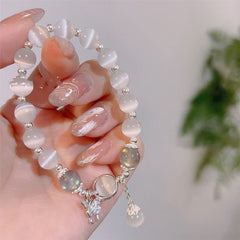 Cat's Eye Moonstone Butterfly Bracelet – White cat's eye beads with silver butterfly charm held in manicured hand, pink marble nails, white and green foliage blur