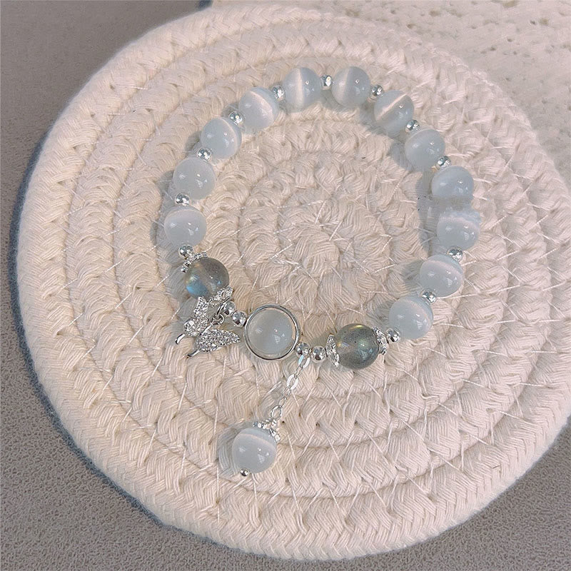 Cat's Eye Moonstone Butterfly Bracelet – Pale blue cat's eye beads with moonstone cabochon and butterfly charm on cream woven circular mat, gray surface top-down