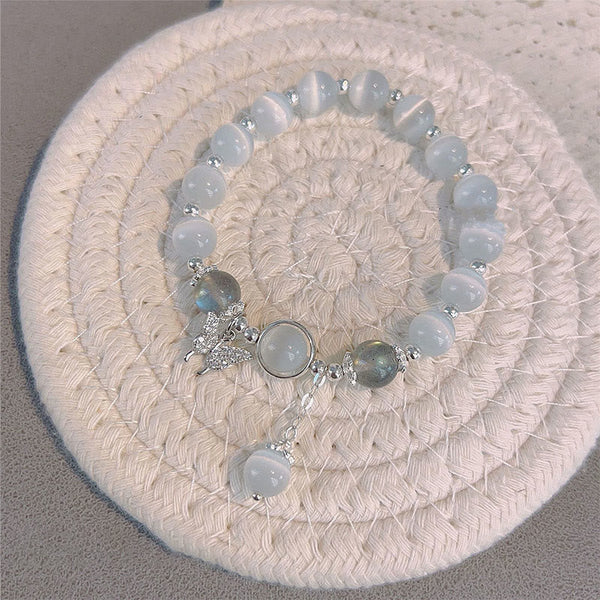 Cat's Eye Moonstone Butterfly Bracelet – Pale blue cat's eye beads with moonstone cabochon and butterfly charm on cream woven circular mat, gray surface top-down