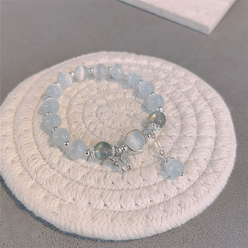 Cat's Eye Moonstone Butterfly Bracelet – Blue cat's eye beads with iridescent labradorite accents and crystal butterfly charm on cream mat, gray surface elevated angle