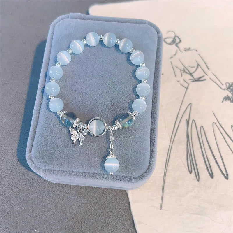 Cat's Eye Moonstone Butterfly Bracelet – Blue cat's eye beads with butterfly crystal charm and dangling pendant on gray-blue velvet with floral sketch paper, overhead