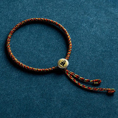 Chinese Zodiac Natal Buddha Bracelet – Red gold green braided cord with brass Sanskrit mantra bead, blue fabric flat lay product shot