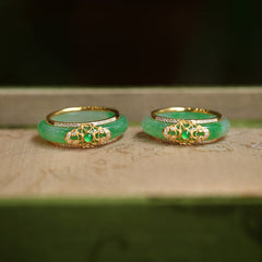18K Gold Green Chalcedony Strength Ring – Two apple-green jade bangles with gold filigree diamond mounts and emerald cabochons on beige fabric, low front angle