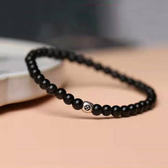 Black Obsidian Smoky Quartz Bracelet – Black glossy beads with silver and gold peace symbol accent bead on peach beige surface, low side angle with bokeh background