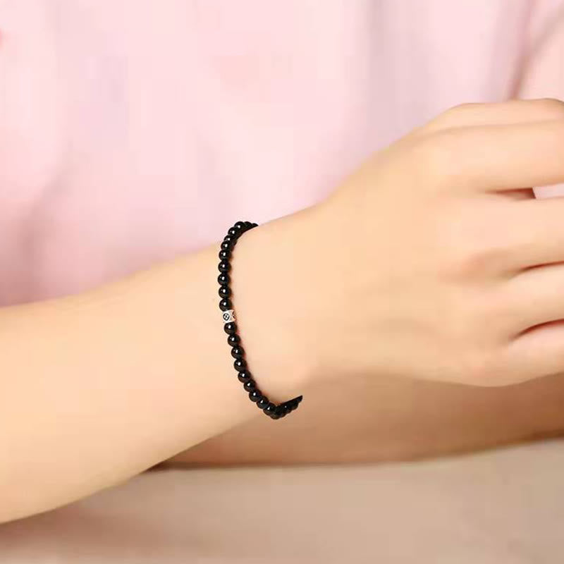 Black Obsidian Smoky Quartz Bracelet – Black beaded bracelet with silver logo accent bead worn on wrist with pink sleeve, warm neutral background side view