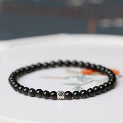 Black Obsidian Smoky Quartz Bracelet – Glossy black beads with silver floral accent bead on white ceramic dish with painted botanical motifs, elevated side angle