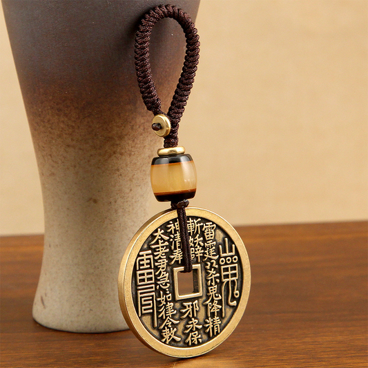 Vajra Knot Shan Gui Keychain – dark brass coin amulet with brown braided cord beside ceramic vase, wooden table, talisman keychain
