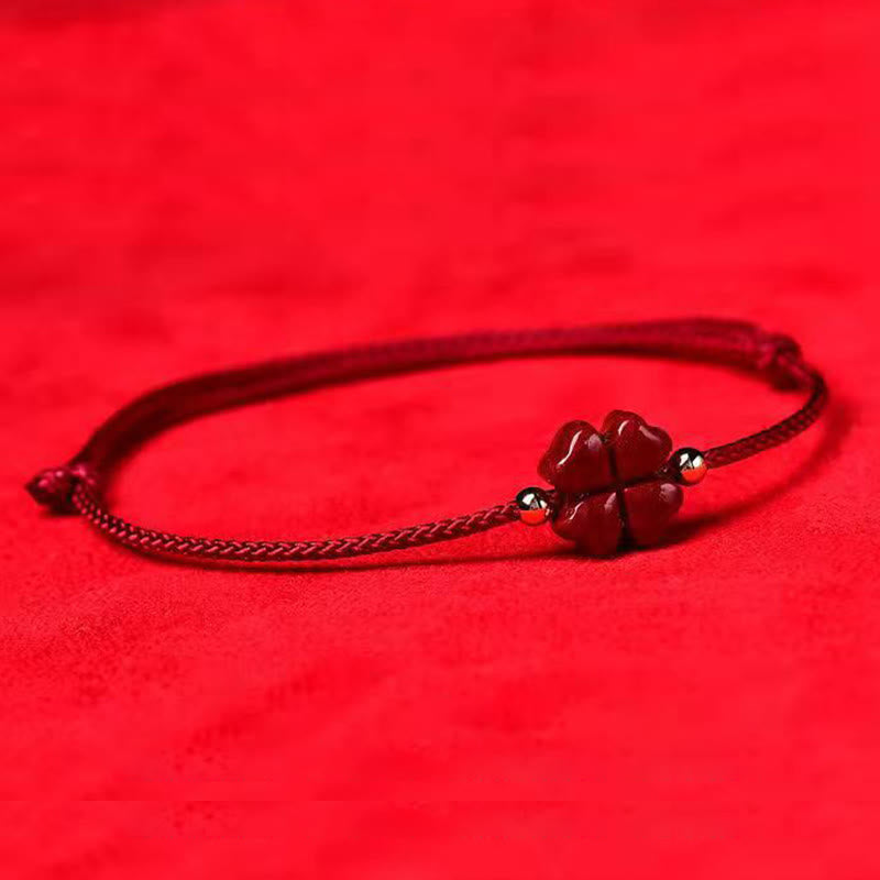 Natural cinnabar four leaf clover red string bracelet with dark red carved charm gold flanking beads and sliding knot closure on deep red textured fabric surface