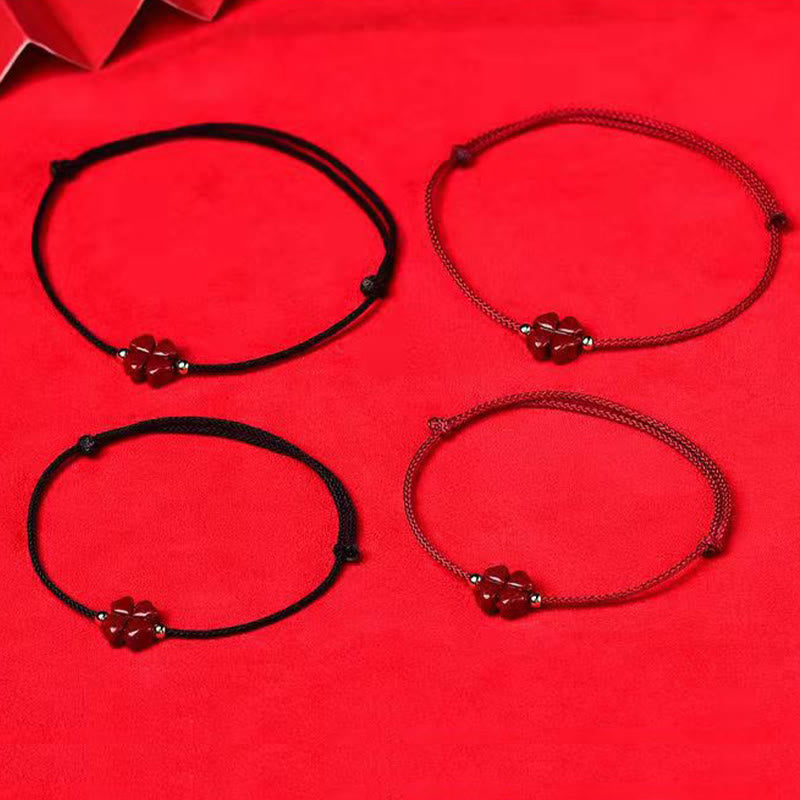Four variant red string bracelets in 2x2 grid showing black cord and burgundy cord options with cinnabar four leaf clover charm on red fabric with red envelope accent