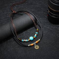 Turquoise Dzi Bead Protection Necklace – close-up showing turquoise bead with natural black matrix veining, Dzi eye beads in cream and brown, amber cylindrical beads and Chinese coin charm on dark braided leather, draped on black fabric with cylindrical display prop