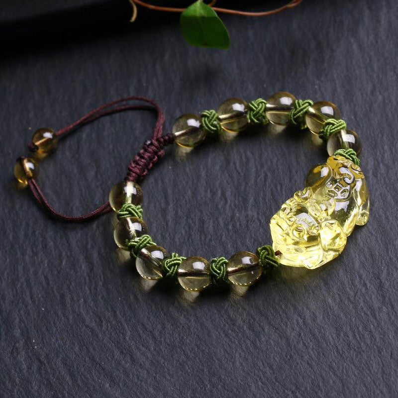 Natural Citrine PiXiu Wealth Bracelet – golden-yellow translucent citrine beads with carved PiXiu wealth charm centerpiece on olive green macrame cord with burgundy adjustable closure, displayed on dark slate surface with natural leaf accents, three-quarter overhead angle