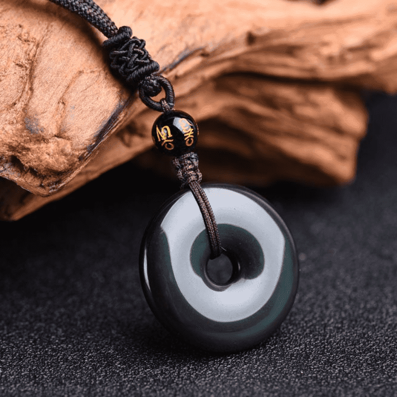 Macro close-up of silver sheen obsidian bi disc pendant necklace showing metallic chatoyant eye effect with gold-character mantra bead on Chinese knotted cord on dark fabric with wood prop