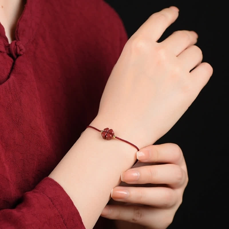 Cinnabar four leaf clover bracelet worn on model wrist with traditional red garment against black background showing red cord carved vermilion charm and gold accent beads