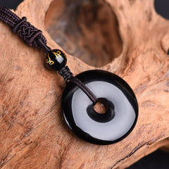 Polished black obsidian bi disc pendant necklace featuring gold Sanskrit mantra accent bead and traditional Chinese knotwork on dark woven cord against rustic wood background