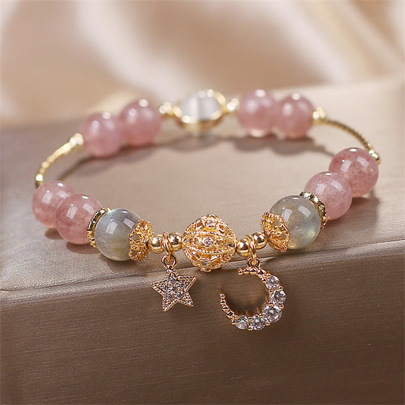 Strawberry Quartz celestial bracelet with translucent pink beads, gray-blue labradorite accents, gold filigree centerpiece, crystal star and moon charms on wood.