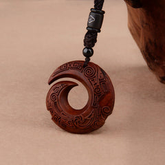Red sandalwood peace buckle pendant necklace showing carved spiral koru design with intricate meander and wave patterns on dark reddish-brown wood with black cord and cloud-motif accent bead