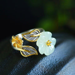 White Jade Plum Blossom Silver Ring - Adjustable open-band ring with carved white jade flower, gold stamen center, and oxidized silver leaves on dark stone surface