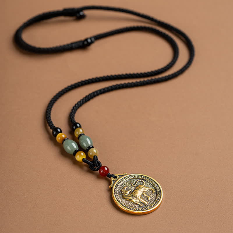 12 Chinese Zodiac Fortune Necklace Pendant – Tiger zodiac medallion necklace flat lay on warm beige background, gold brass circular pendant with walking tiger relief, Greek key border pattern, black cord with green jade and amber stone beads, cultural heritage jewelry